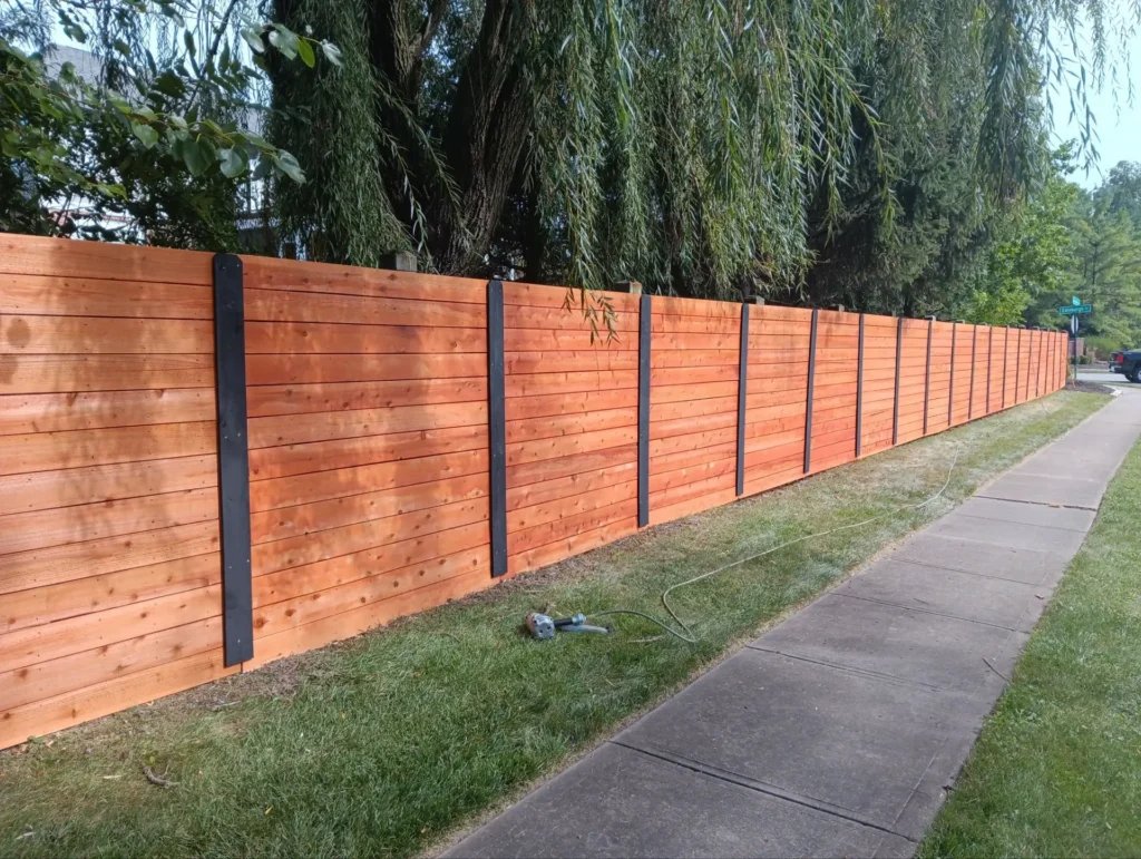 Long stretch of newly installed horizontal wooden fencing with black metal posts along a residential sidewalk, surrounded by lush greenery and overhanging tree branches.